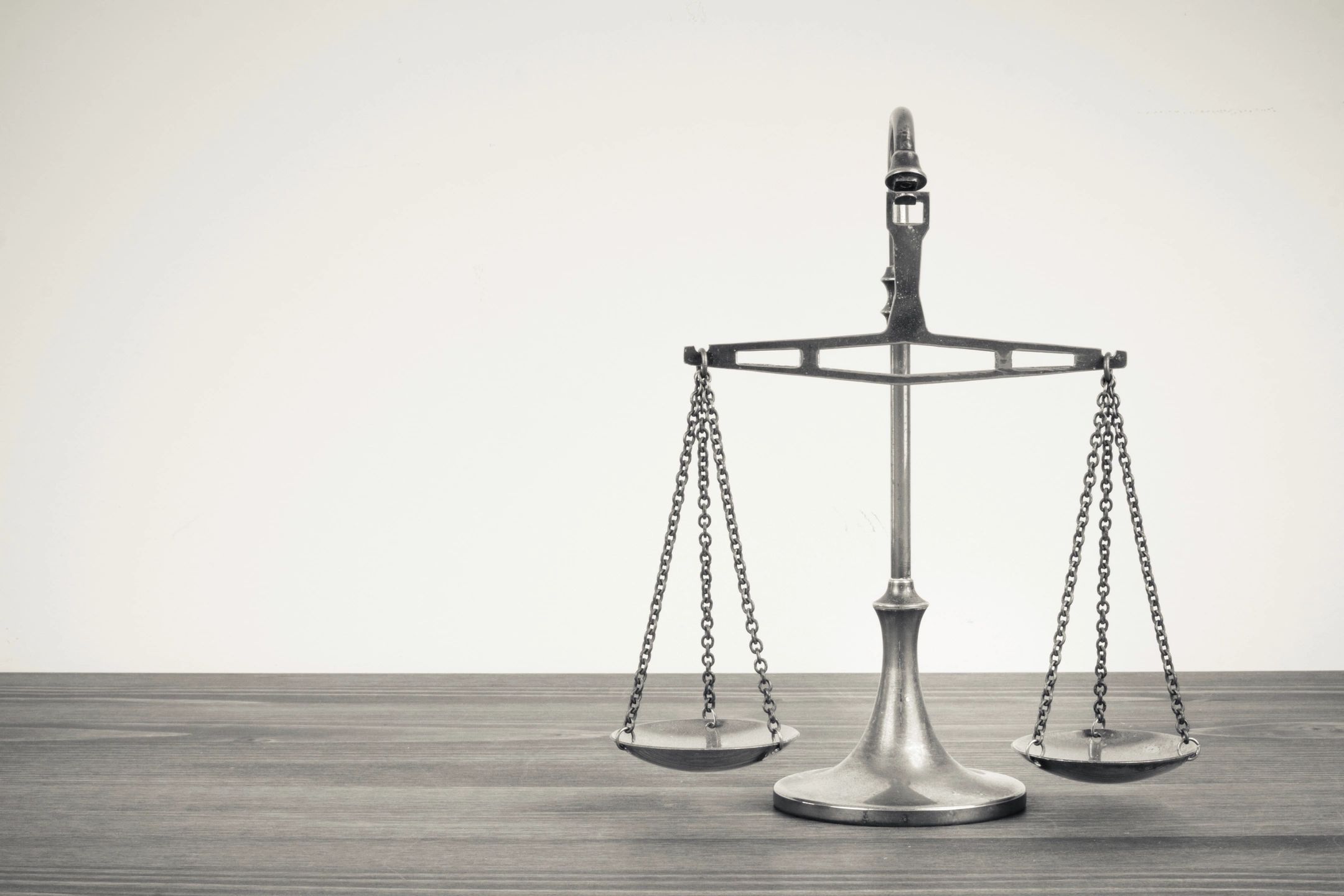 Black and white photo of a balance scale standing on a wooden table against a plain light background, minimalist and formal.