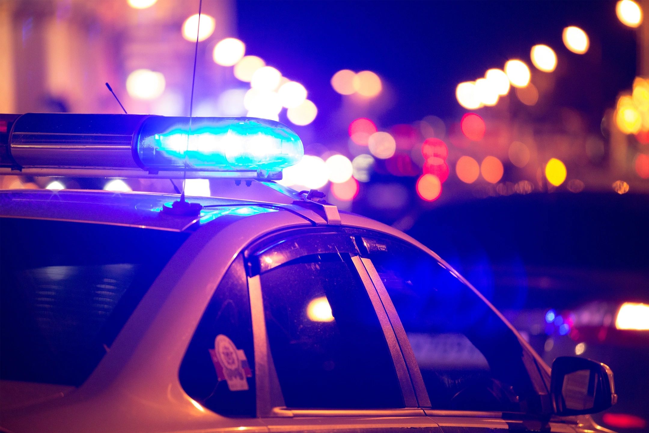Night scene of a vehicle roof with an illuminated blue emergency light bar, city lights blurred in the background, dark and urgent mood.