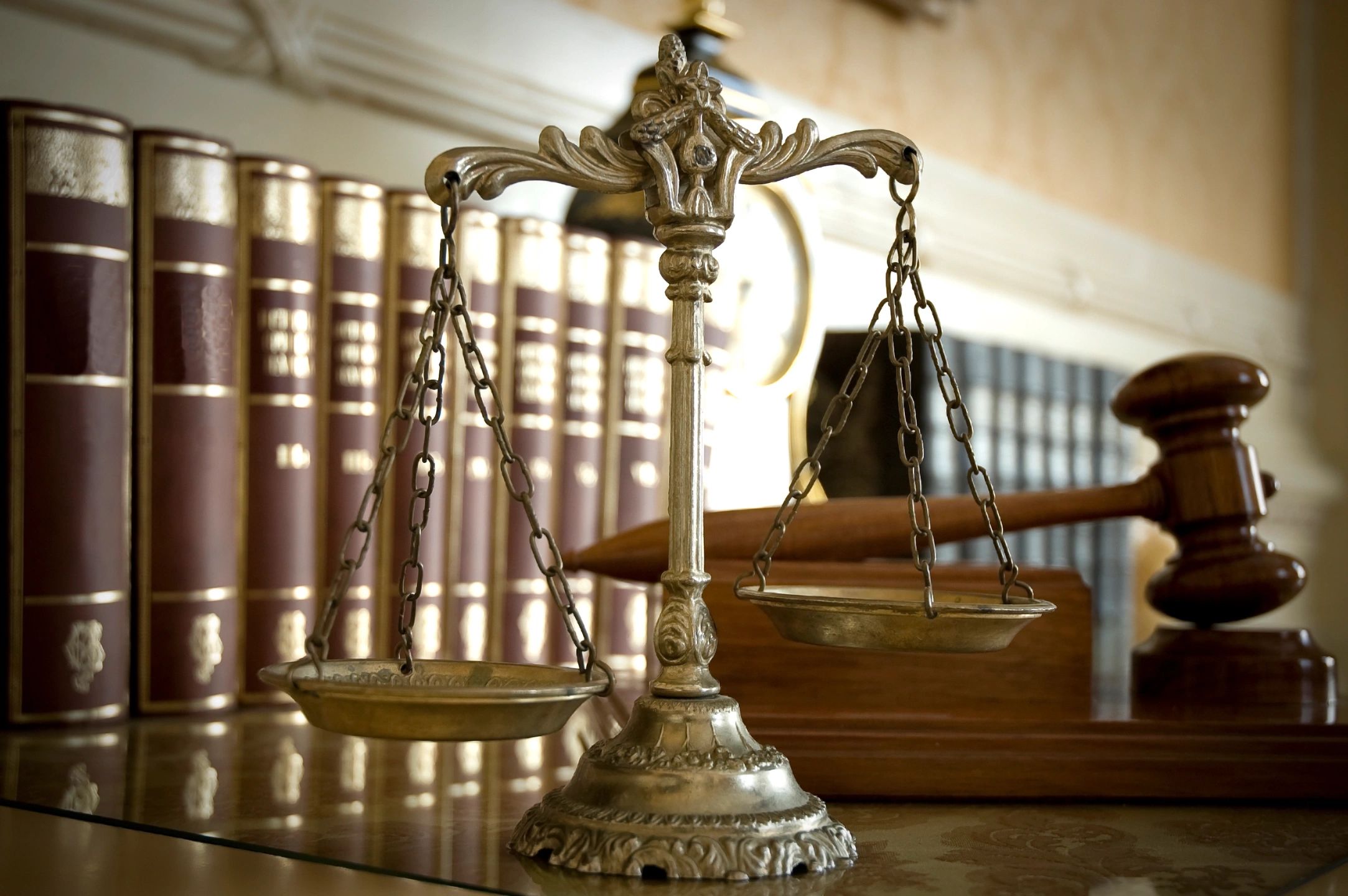 Close-up of a brass balance scale on a desk with a wooden gavel and rows of leather-bound books in the background, warm amber and brown tones, dignified mood.